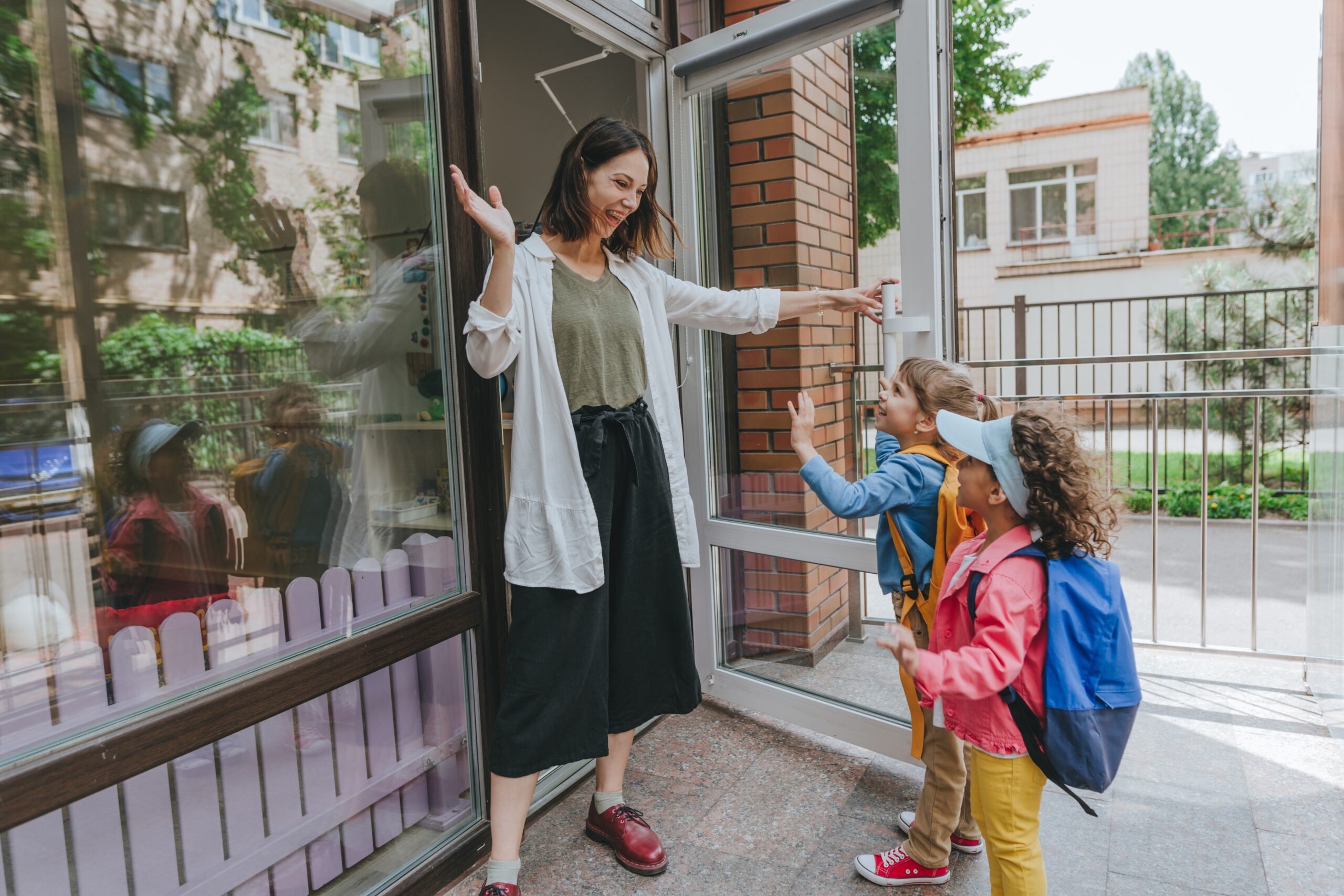A woman at the front door of a daycare welcoming two children.