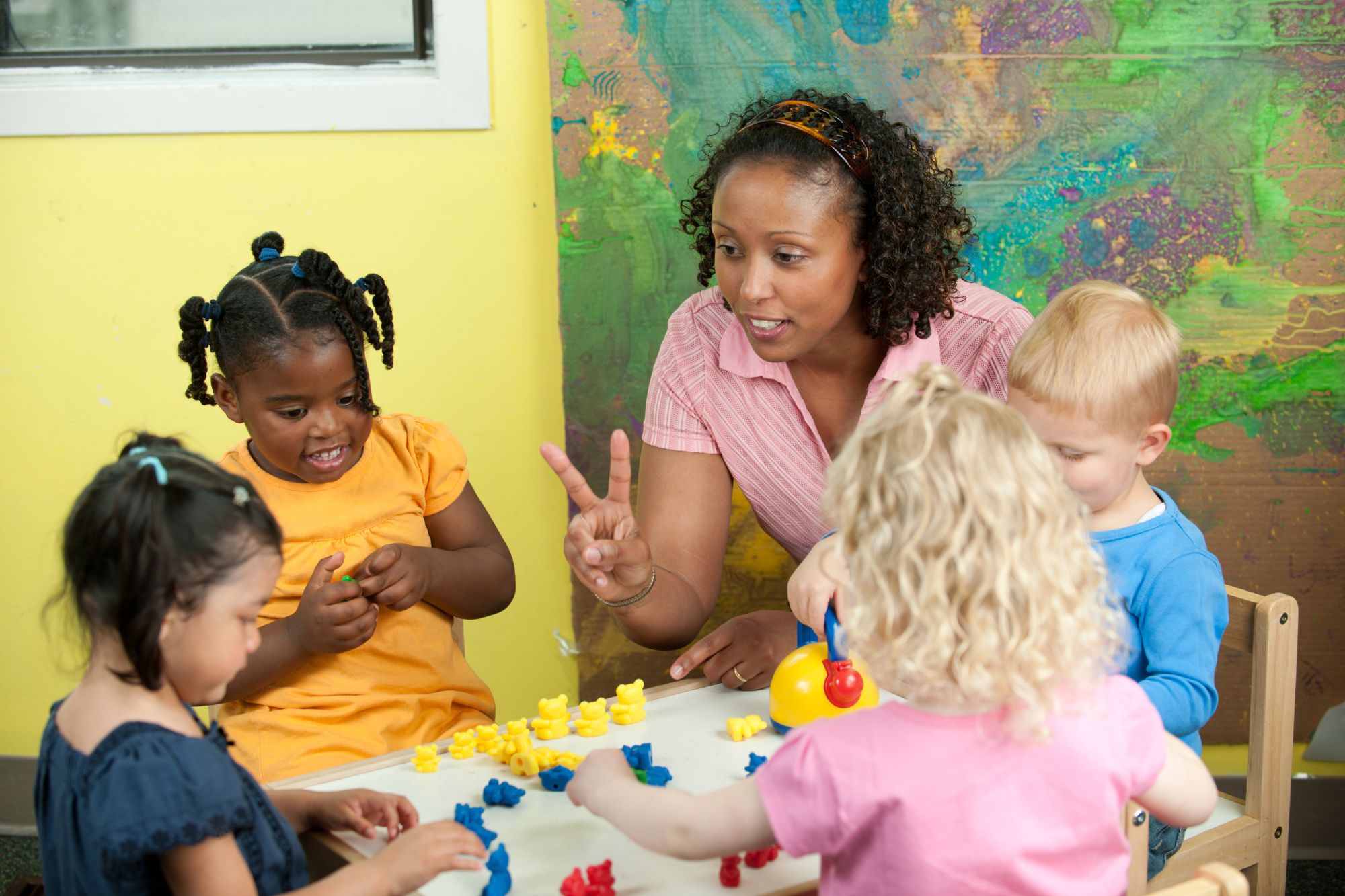 Child care provider sits at small circular classroom table with four young children and teaches them to count