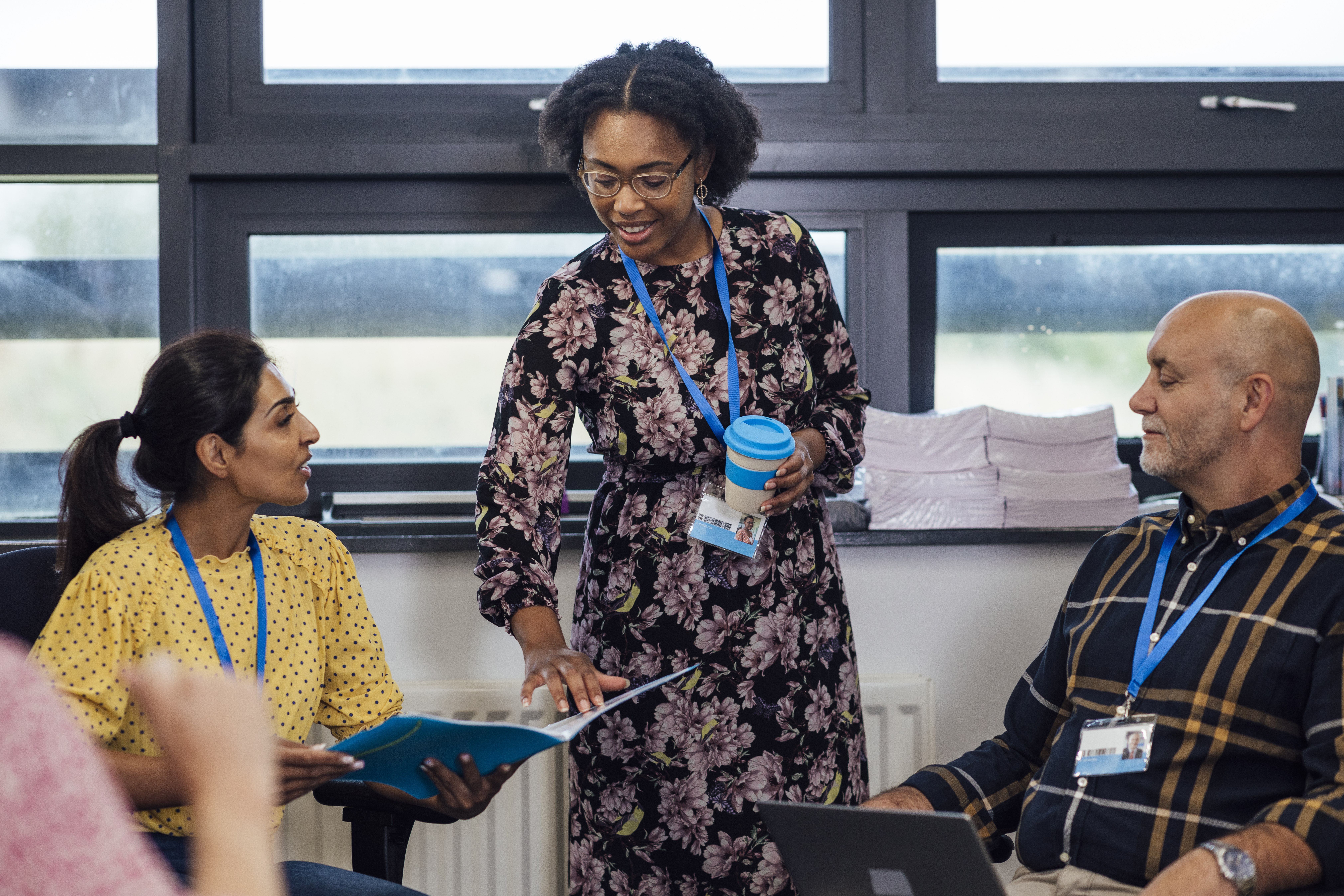 A small group of teachers in a meeting, discussing work on a laptop and in a folder, wearing smart-casual clothing. They are in an office within a secondary school.