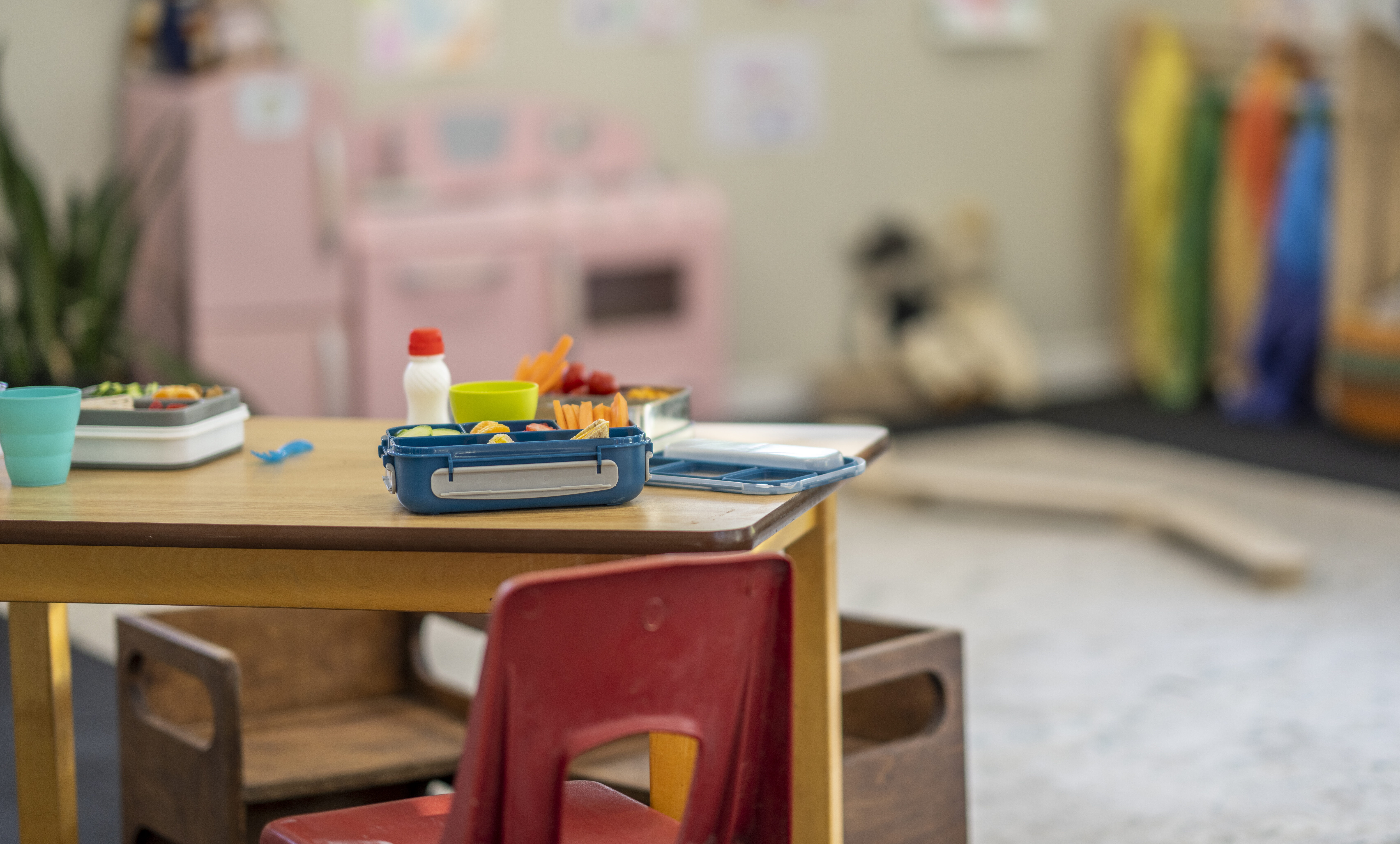 A daycare classroom sits empty of children as they take a break to play outside. A table with children's lunch boxes sitting out is seen in the foreground.