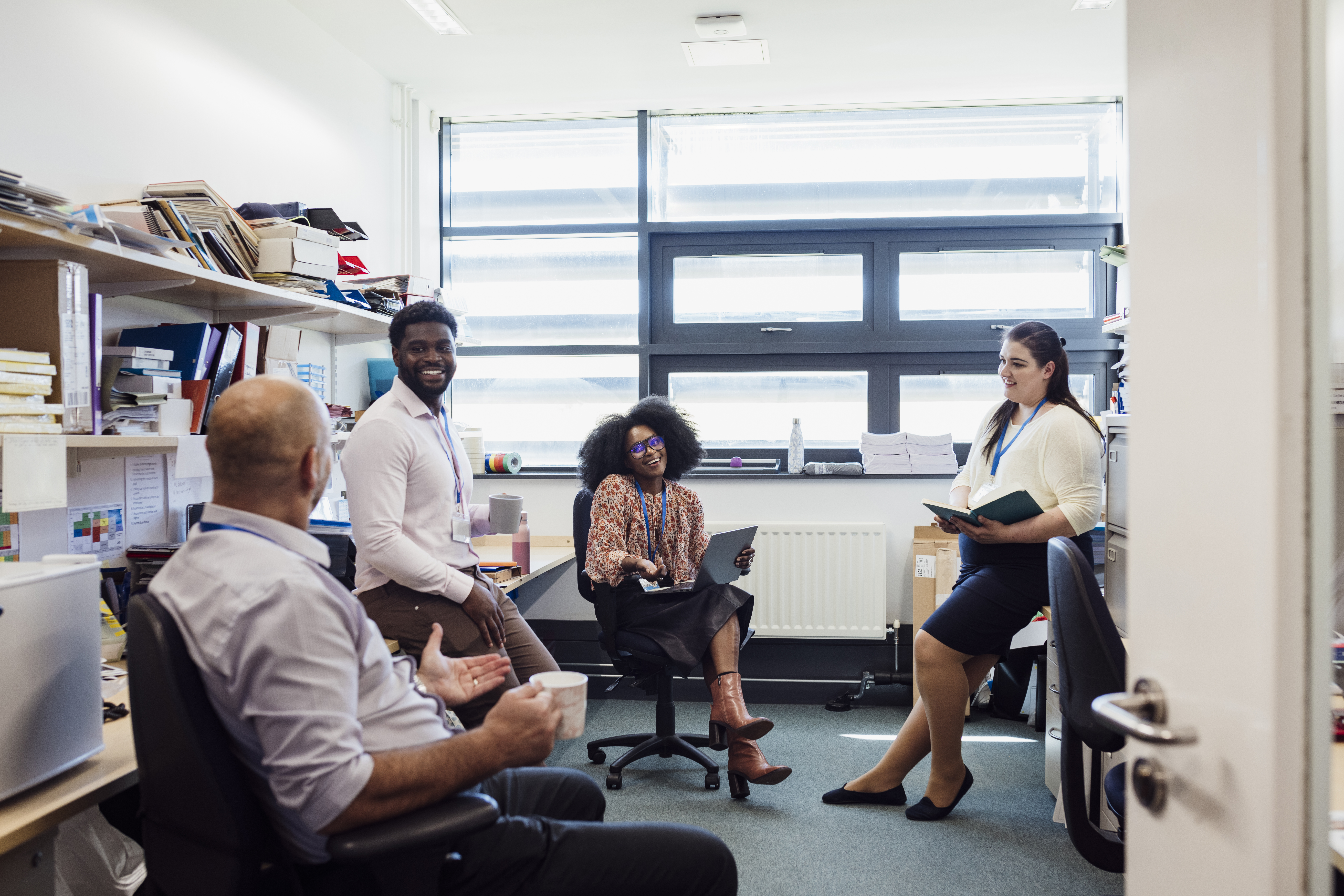 Teachers in an office together in the school that they work at. They are taking a break from teaching and bonding with each other while they discuss lessons.