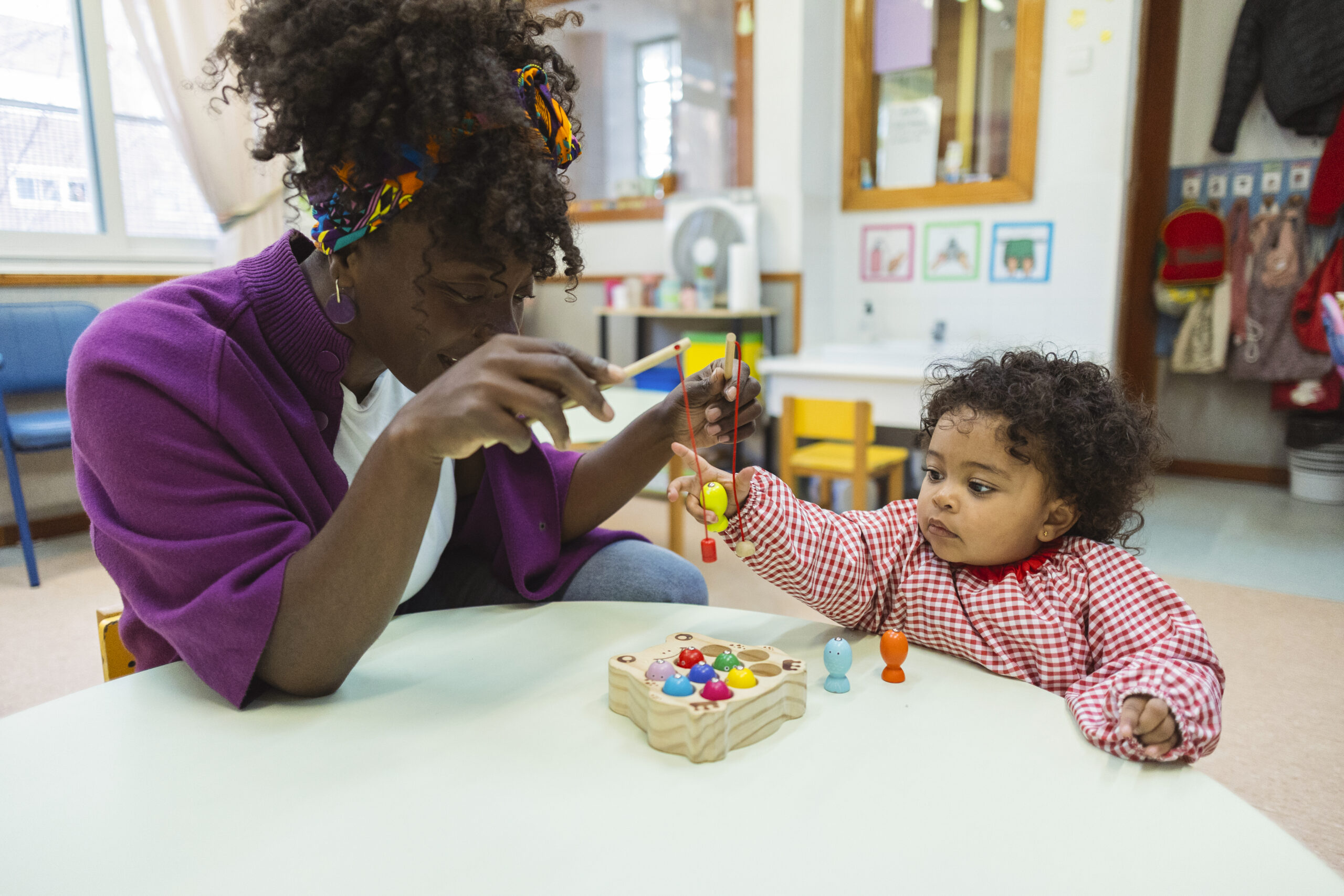 African american teacher engaging with a young toddler, who is focusing on a colorful wooden fishing game, promoting early childhood education and development in a bright daycare setting